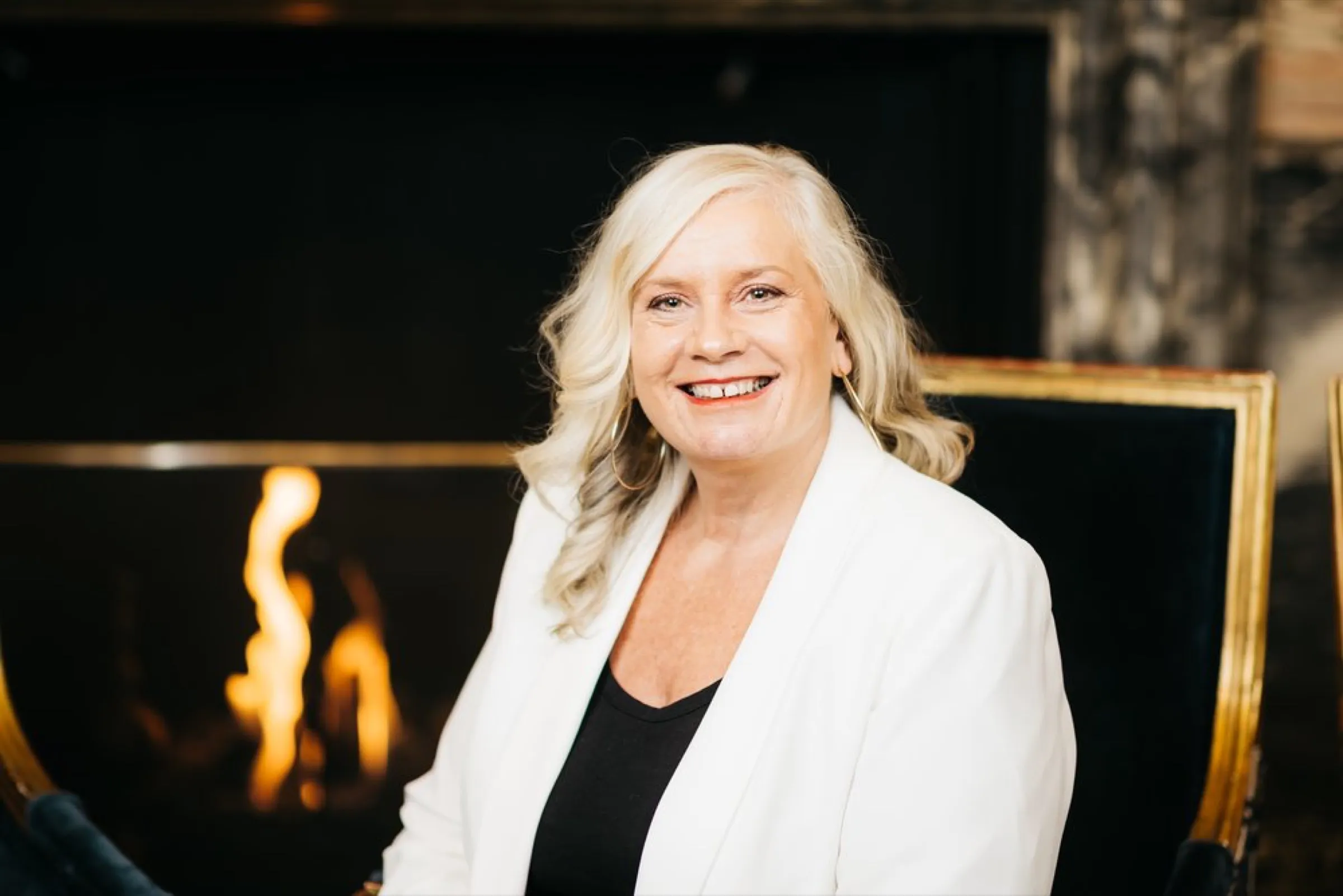 Dr Alison Edgar MBE, seated by a fireplace in a white blazer.