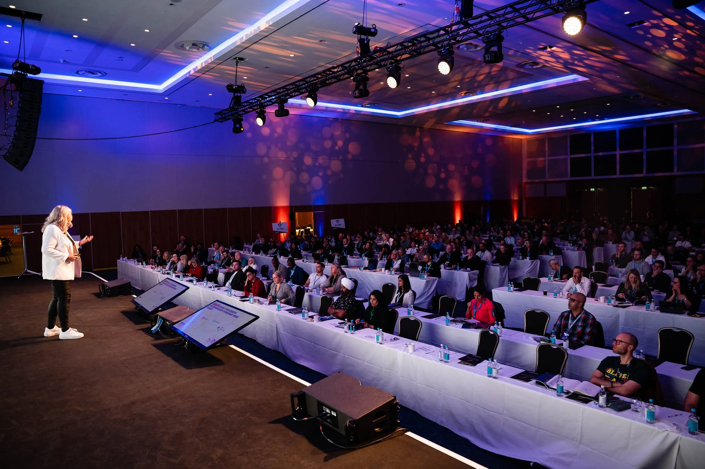 Dr Alison Edgar MBE on stage, speaking to a packed conference room of delegates seated at long tables.