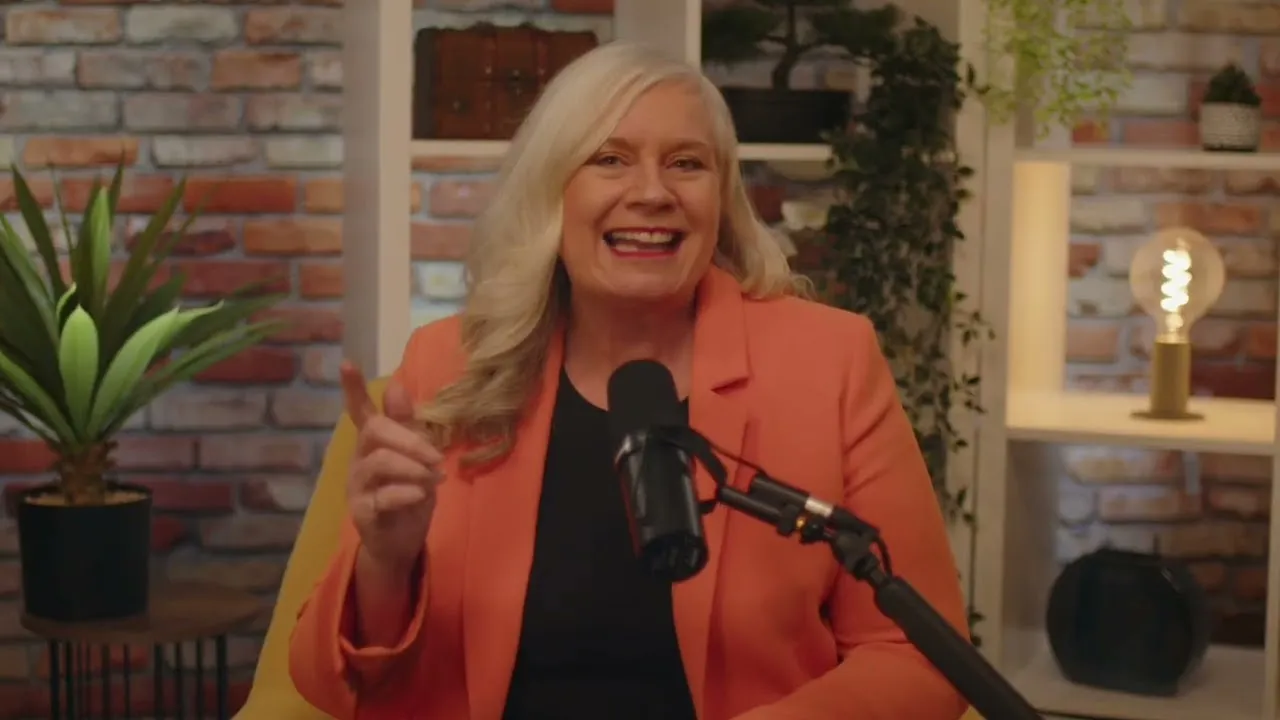 Dr Alison Edgar MBE at a microphone, mid-explanation, in a coral jacket.